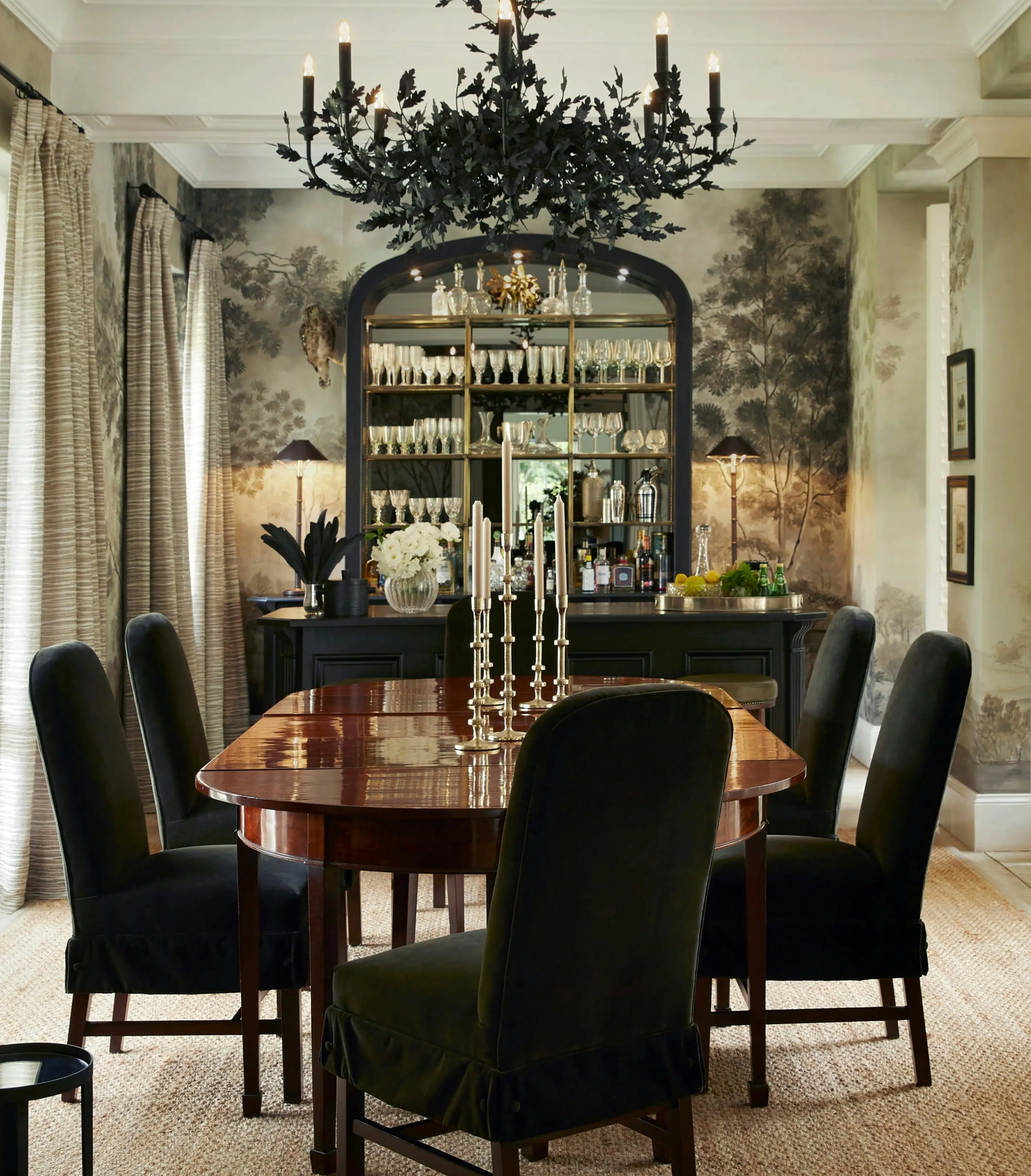 A polished wood dining table holds metal candlesticks and is surrounded by six chairs in a dark fabric. In the background, a tall mirrored sideboard holds stemware, vases and a pair of lamps.