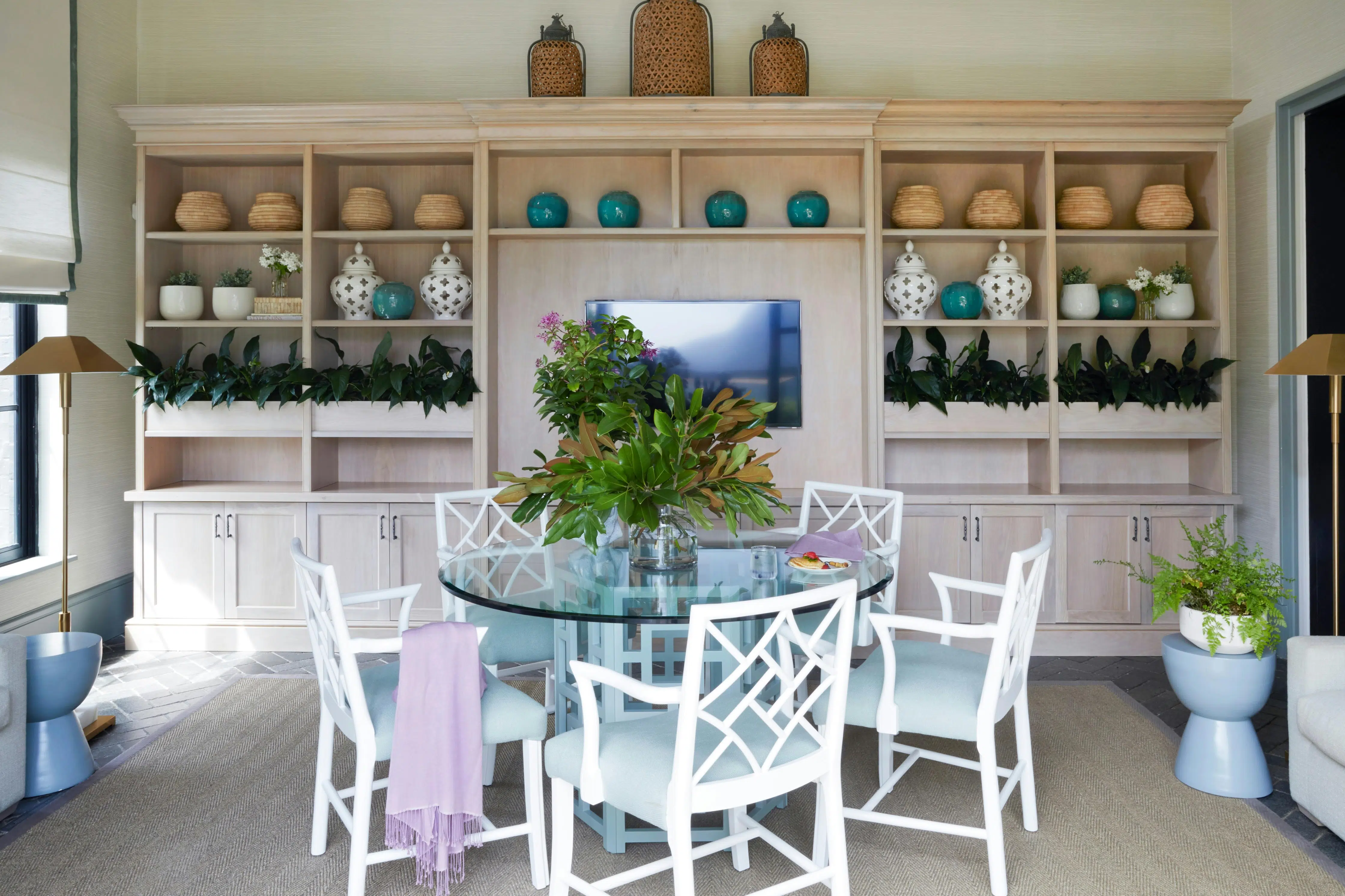 White-framed chairs and a central, glass-topped table holding a large vase of mixed greenery all sit in front of a wall of shelves holding plants and ceramic vessels