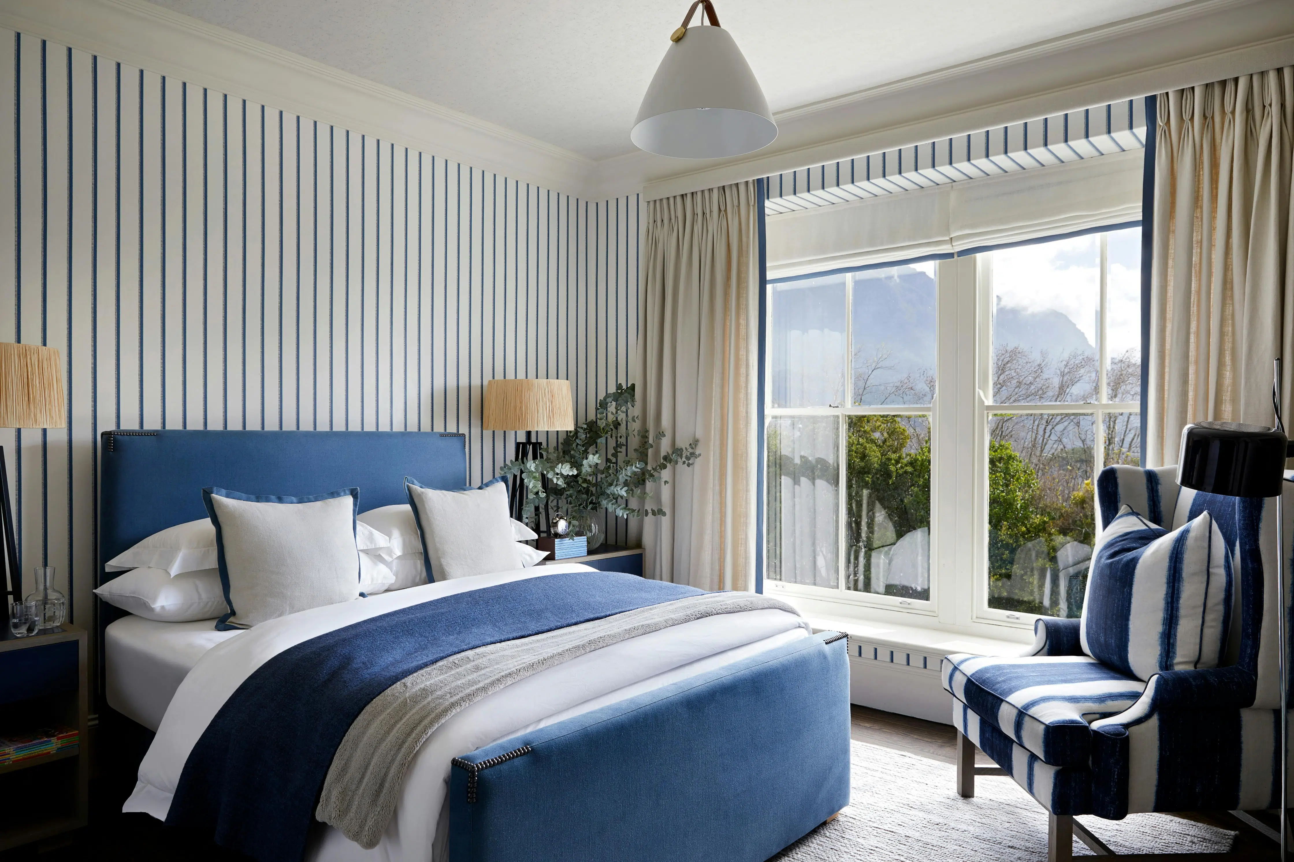 A contemporary bedroom with blue and white striped wallpaper, a navy blue bed layered in blue, white and grey bedding and an armchair in the corner next to a window with a view of trees.