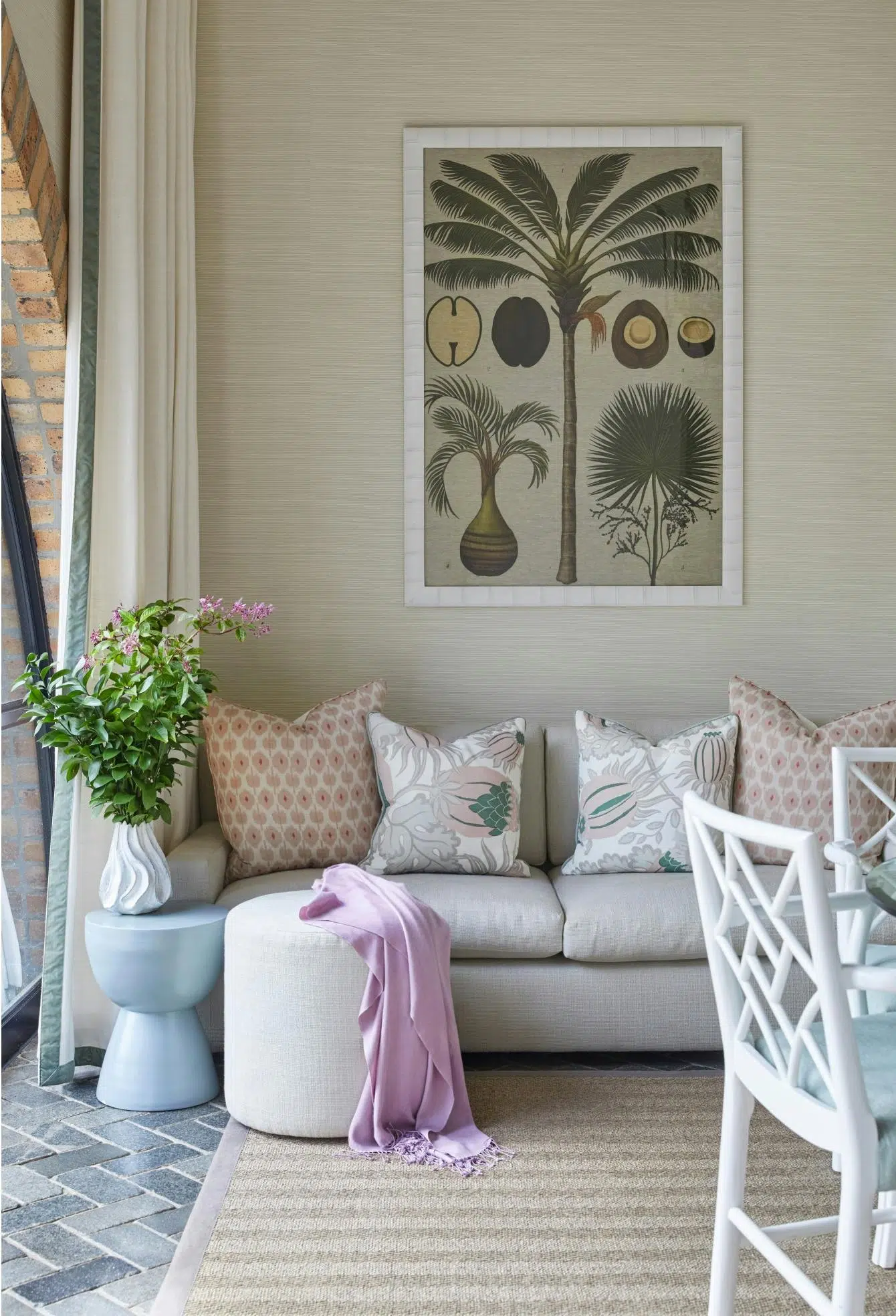 A botanical illustration of a palm and its fruits hangs on the wall behind a beige couch holding patterned cushions in pinks, corals and greens. In front of the couch are a contemporary blue side table holding a vase that contains greenery and flowers, a pouffe with a lilac throw draped over it, and a grass carpet on a dark brick floor